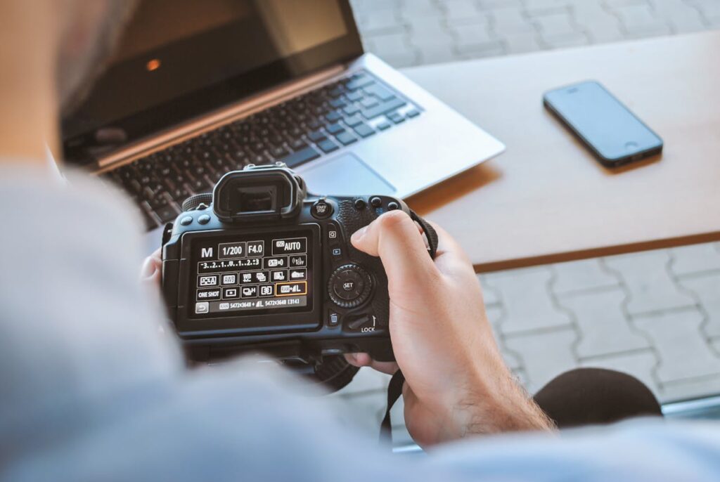 Close-up of hands holding DSLR camera at a workspace with laptop and smartphone.